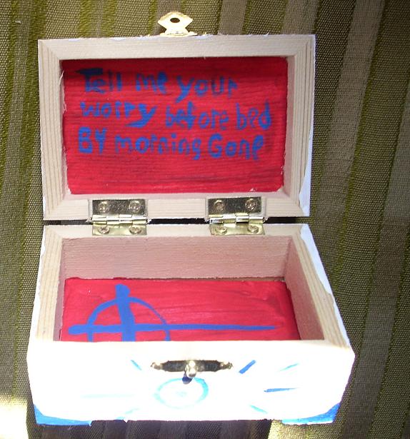 这是一个手工制作的木制烦恼盒，打开的盒子的内盖上用蓝色油漆写着“睡前告诉我你的烦恼。”到早上就没了。”盒子的内部是红色的，一个蓝色的十字画在盒子的底部。