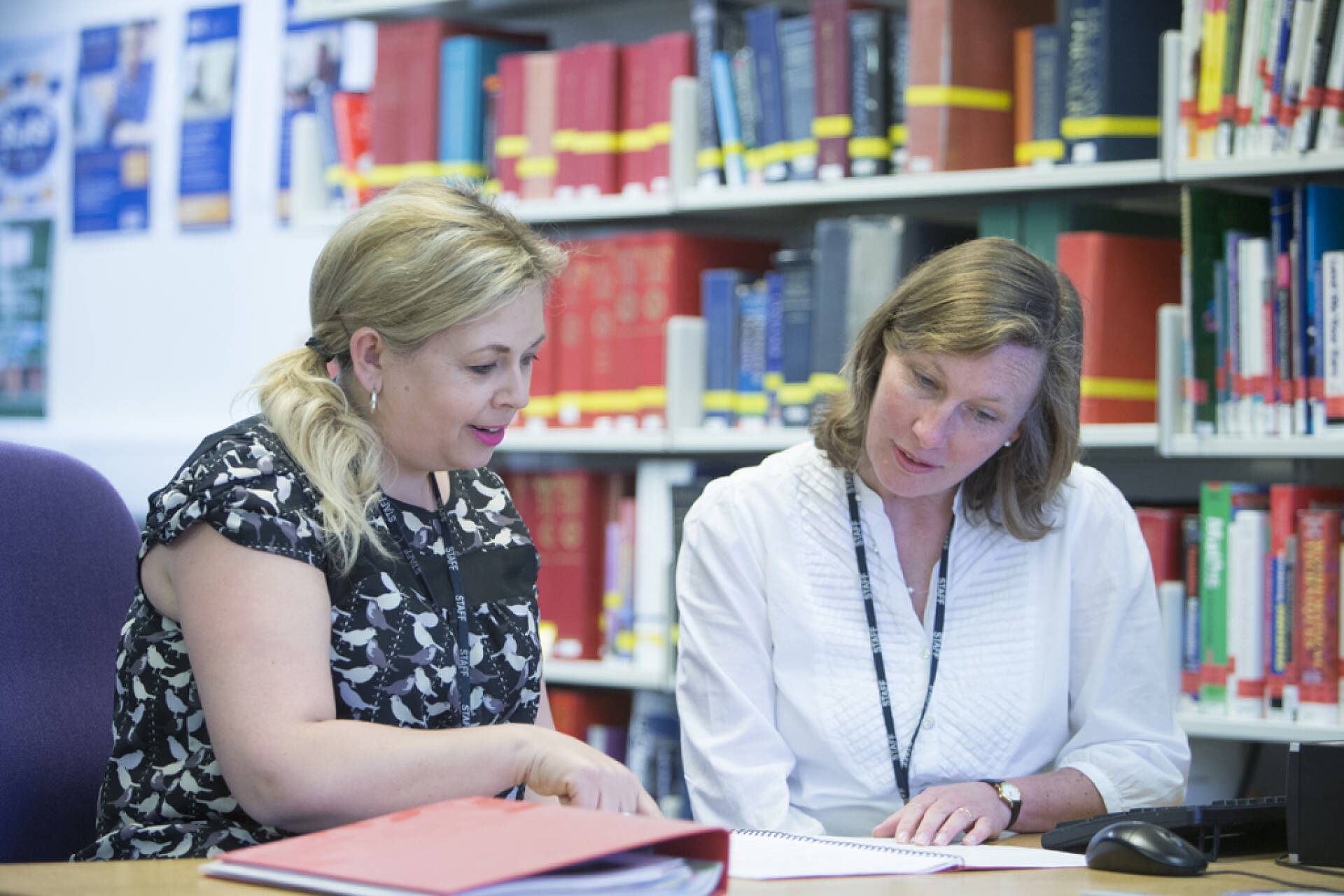 两名女同事在图书馆书架上的书的背景下，在笔记本上学习笔记