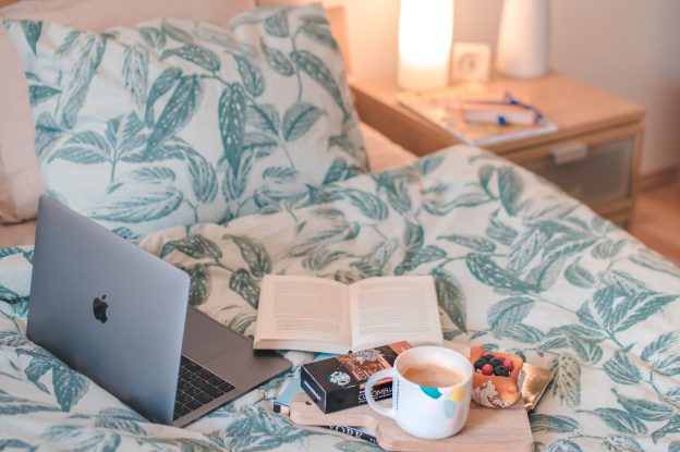 笔记本电脑，一杯茶，打开的书和一份水果糕点都放在一张铺好的床上，床边有一个床头柜。
